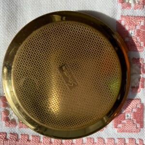 May include: A round, gold-toned Stratton compact with a textured, woven-like surface. The compact has a shiny, polished finish and the word "Stratton" is subtly engraved on the surface. The compact is a vintage beauty accessory.