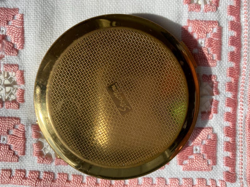 May include: A round, gold-toned Stratton compact with a textured, woven-like surface. The compact has a shiny, polished finish and the word "Stratton" is subtly engraved on the surface. The compact is a vintage beauty accessory.