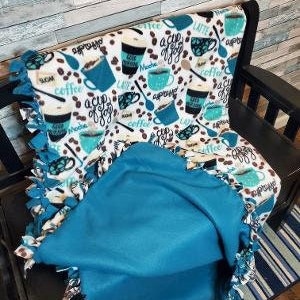 May include: A fleece blanket with a coffee-themed pattern in shades of teal, black, and white, draped over a chair. The design includes coffee cups, beans, and the words "a cup of joy" and "coffee". The blanket has a teal border.