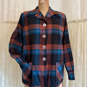 May include: A vintage plaid jacket in black, teal, and orange, displayed on a mannequin. The jacket features a collared neckline, large white buttons, and two front pockets. The plaid pattern is a classic design.
