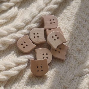 Puede incluir: Un conjunto de botones de madera en forma de gato, de color marrón claro, con cuatro agujeros cada uno, dispersos sobre una tela de punto crema texturizada. La tela presenta un diseño de punto trenzado.