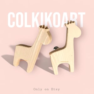 May include: Two wooden giraffe-shaped cabinet knobs. The knobs are light-coloured wood with a natural grain. The image includes the text "COLKIKOART" and "Only on Etsy" on a pink background.
