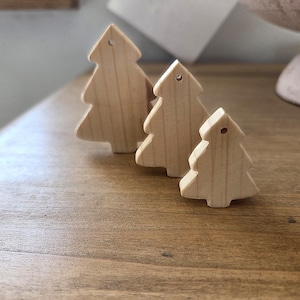 Könnte beinhalten: Drei helle Holzanhänger in Tannenbaumform in verschiedenen Größen, jeweils mit einer einfachen Silhouette und einem kleinen Loch an der Spitze zum Aufhängen. Die natürliche Holzmaserung ist auf den glatten, flachen Oberflächen dieser drei Baumfiguren sichtbar.