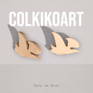 Può includere: Due ornamenti in legno a forma di uccello con finitura in legno naturale. Gli ornamenti sono esposti su uno sfondo grigio chiaro. Il testo "COLKIKOART" è in alto e "Only on Etsy" in basso.