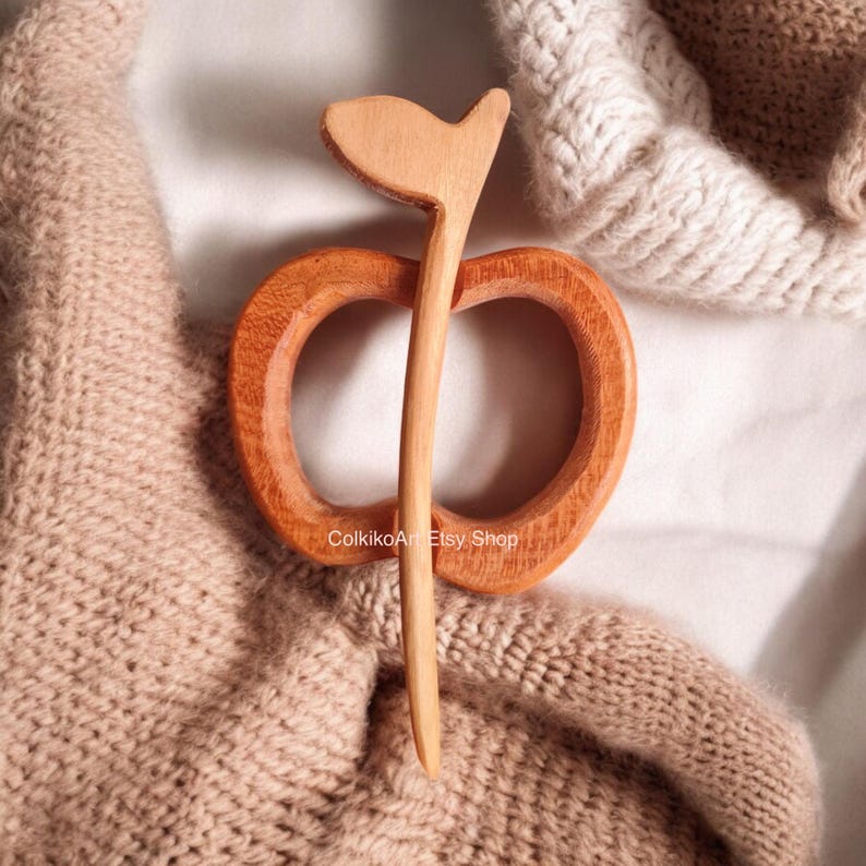 May include: A wooden apple-shaped hair clip with a long, thin stem. The clip is made of natural wood and has a light brown color. The clip is lying on a brown knitted fabric.