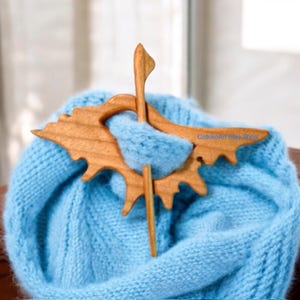 May include: A wooden shawl pin in the shape of a bird with a natural wood finish. The pin is holding a light blue knitted scarf.