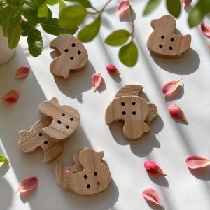 Op de afbeelding: Een verzameling houten eendvormige knopen met vier kleine gaatjes, verspreid op een wit oppervlak. De knopen zijn lichtbruin en omgeven door roze bloemblaadjes en groene bladeren.