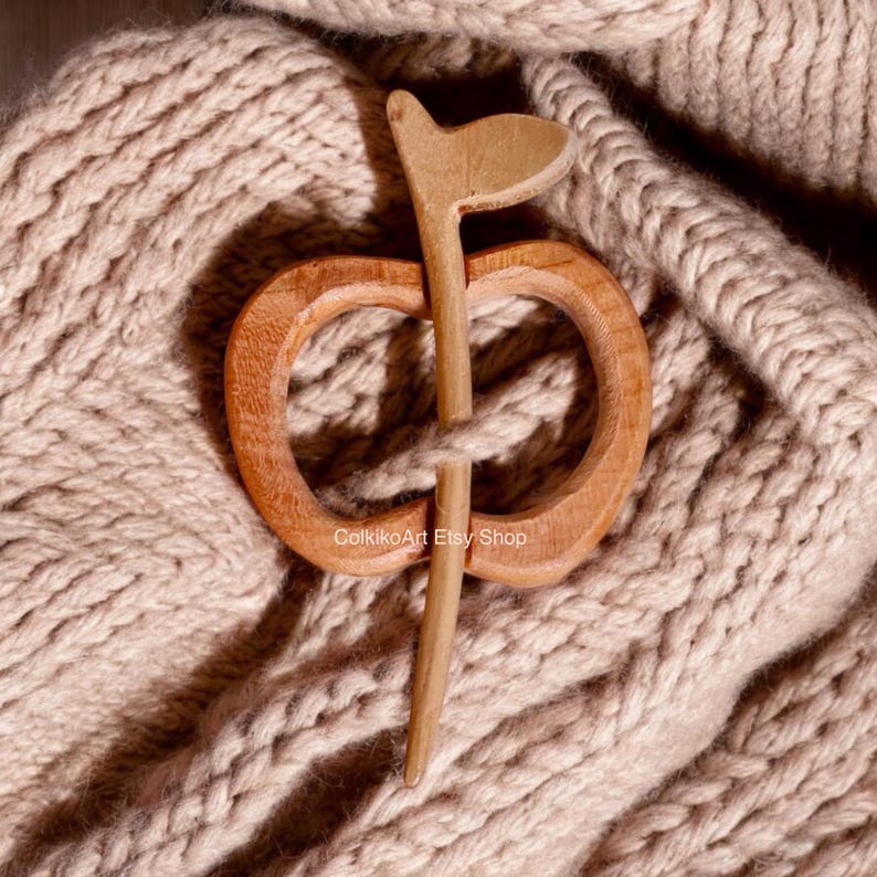May include: A wooden apple-shaped shawl pin with a long, thin stem. The pin is resting on a beige knitted fabric.