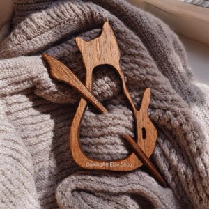 Könnte beinhalten: Eine schalkhafte Holzschalspange in Katzenform mit einem langen, dünnen Holzstab, der durch den Körper der Katze geht. Die Spange liegt auf einem braunen Strickstoff.