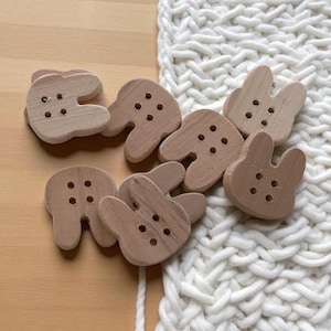 Peut inclure: Un assortiment de boutons en bois en forme de dents et de lapins. Les boutons sont de couleur marron clair avec trois ou quatre trous pour la couture. Ils sont disposés sur une surface en bois clair à côté d'un tissu tricoté blanc.