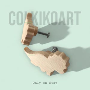 May include: Two wooden dinosaur-shaped cabinet knobs with metal screws. The knobs are light brown and have a textured surface. The background is a light teal colour. The text "COKIKOART" is at the top and "Only on Etsy" at the bottom.
