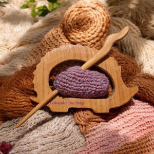 May include: Wooden hedgehog shawl pin with a purple knitted scarf wrapped around it. The pin is made of natural wood and has a smooth finish. The scarf is soft and cosy, and the pin is a great way to keep it in place.
