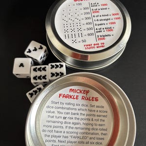 May include: A Mickey Farkle dice game set featuring two round silver metal tins with printed rule and scoring guides. The game includes six white six-sided dice with black Mickey Mouse head icons replacing the standard pips on each face.
