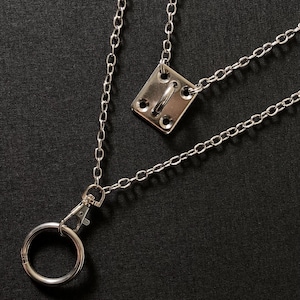 May include: A silver-toned metal lanyard featuring a rectangular plate with four corner screw holes and a central loop. The chain connects to a lobster claw clasp that holds a circular ring. The hardware has a polished finish and metallic sheen.