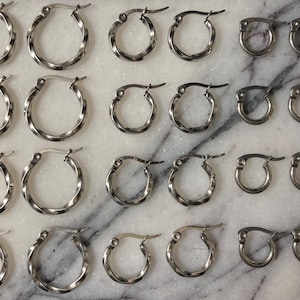 May include: An assortment of silver-colored hoop earrings in various sizes. Some hoops have a twisted design, while others are smooth. The earrings are arranged on a white and gray marble surface.