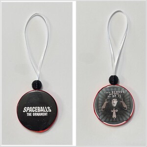 May include: Two round Spaceballs-themed ornaments. One is black with white text that reads "SPACEBALLS THE ORNAMENT". The other has a black and white image of a character with the text "I SAID ACROSS HER NOSE, NOT UP IT."
