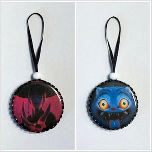 May include: Two round ornaments with black trim and black ribbons. One features a figure in red and black, the other a blue cat with yellow eyes and sharp teeth. Each has a white bead.