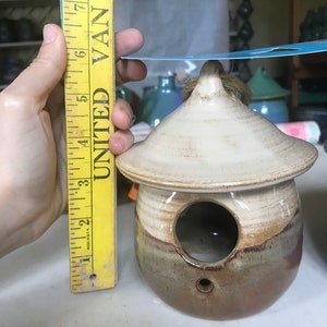 May include: A small, brown and white ceramic birdhouse with a round opening. The birdhouse is about 5 inches tall and has a small hole on the bottom for drainage.