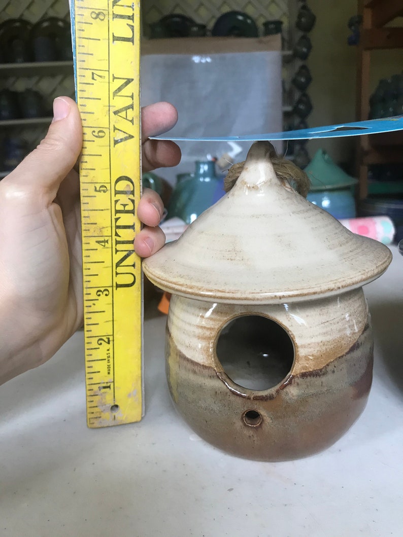 May include: A small, brown and white ceramic birdhouse with a round opening. The birdhouse is about 5 inches tall and has a small hole on the bottom for drainage.