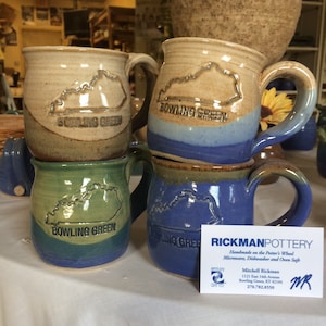 May include: Four handmade ceramic mugs with a blue, green, and brown glaze. Each mug features a raised outline of the state of Kentucky with the words "Bowling Green" printed below. A business card for Rickman Pottery is in the foreground.