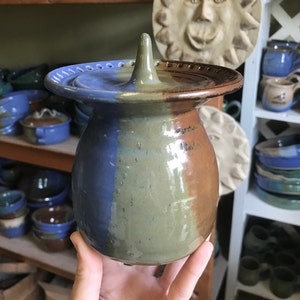 May include: A ceramic jar with a lid, featuring a blue, green, and brown glaze. The lid has a pointed top and a decorative rim.