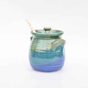 May include: A blue and green ceramic honey pot with a wooden dipper. The pot has a lid and two handles.