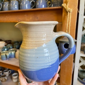 May include: A blue and white ceramic pitcher with a rounded body and a handle. The pitcher has a light blue glaze on the bottom half and a white glaze on the top half. The glaze has a subtle striped pattern.