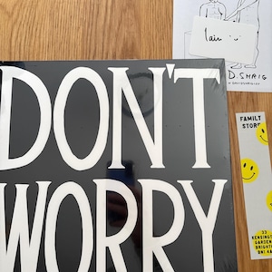May include: A black square vinyl record sleeve with the white text DON'T WORRY printed in large capital letters. Beside it is a white card with a black line drawing of two figures and the text SUPPORT SMALL BUSINESS and D. SHRIG, plus a yellow sticker.