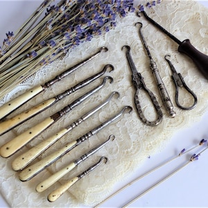 Puede incluir: Una colección de calzadores vintage con mango de hueso. Los calzadores están hechos de metal y tienen una variedad de diseños. Los calzadores se muestran sobre una tela de encaje blanca con ramitas de lavanda secas.