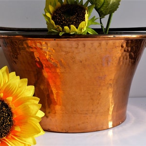 Könnte beinhalten: Ein gehämmerter Kupfer-Eimer mit zwei Eisenhenkeln. Der Eimer ist leer und hat eine glänzende, reflektierende Oberfläche. Eine gelbe Sonnenblume befindet sich im Vordergrund.