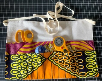 Könnte beinhalten: Ein weißes, zusammenrollbares Nähset aus Canvas mit einem Futter aus gemustertem Stoff in Pink, Gelb und Schwarz. Das Set enthält eine gelbe Schere, ein blaues Maßband und einen gelben Rollschneider.