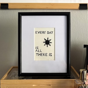 May include: A framed print with a black frame and white mat. The print features the text "EVERY DAY IS ALL THERE IS" with a black sun graphic. The artwork is displayed on a wooden shelf.