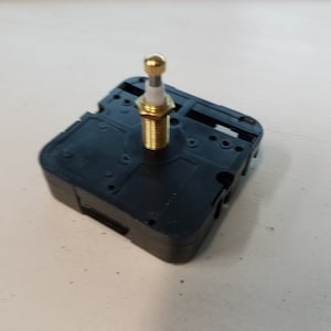 May include: A black clock mechanism with a gold-colored winding stem. The mechanism has a square shape and is designed for use in a clock.
