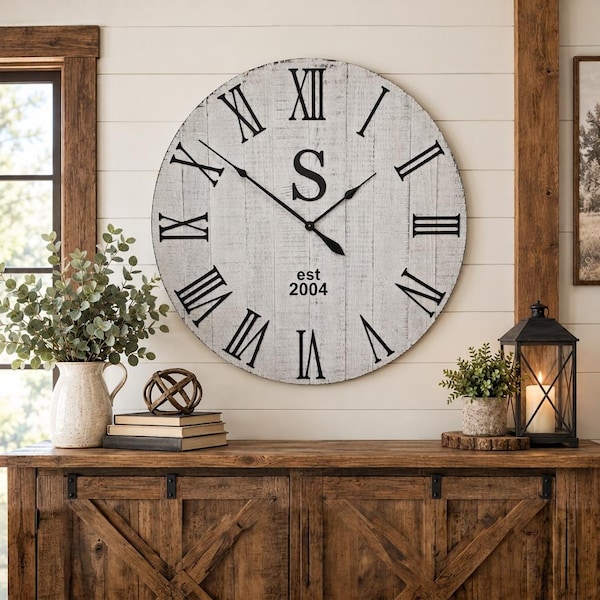 Könnte beinhalten: Eine große runde Wanduhr mit weißem, abgenutztem Holzfinish, schwarzen römischen Ziffern und schwarzen Metallzeigern. In der Mitte befindet sich ein großer schwarzer Buchstabe S über dem Text est 2004. Die Uhr hat einen kreisförmigen Rahmen.