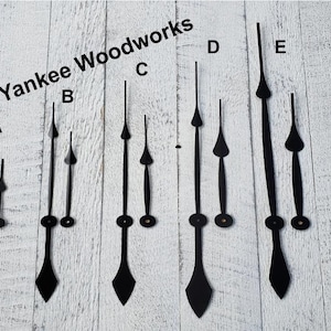 May include: Five sets of clock hands in various sizes, labeled A through E. The hands are black metal with a pointed tip and a round base. The hands are arranged on a white wooden background.