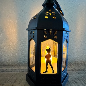 May include: Black metal lantern with a silhouette of Peter Pan inside. The lantern has a candle inside and is lit, casting a warm glow.