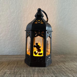 May include: Black metal lantern with a Mickey Mouse silhouette. The lantern has a handle and decorative cutouts. The interior glows with a warm, yellow light, creating a cozy ambiance. A charming decorative piece.
