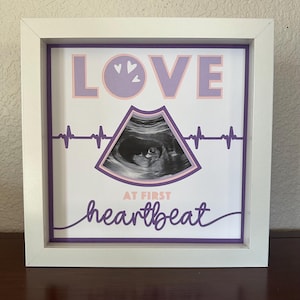 Puede incluir: Obra de arte enmarcada en blanco con la palabra "LOVE" en lavanda, con una imagen de ultrasonido. La frase "AT FIRST heartbeat" está escrita en cursiva debajo. Se incluye un diseño de línea de latido púrpura.