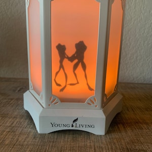 May include: White ceramic diffuser with a frosted glass insert featuring a silhouette of two frogs. The diffuser has a decorative design and the words "Young Living" on the base.