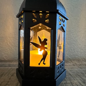 May include: Black metal lantern with a cutout of Tinkerbell inside. The lantern has a candle inside, casting a warm glow.