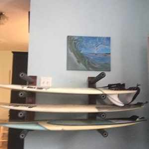 May include: Three surfboards are mounted on a wall rack. The surfboards are stacked on top of each other. The top surfboard is white, the middle surfboard is white, and the bottom surfboard is blue and white.