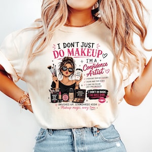 May include: A cream-colored t-shirt featuring a graphic illustration of a person with makeup brushes, a coffee cup, and cosmetics. The text reads: I DON'T JUST DO MAKEUP I'M A Confidence Artist, I HIGHLIGHT YOUR BEST FEATURES, I BLEND AWAY YOUR DOUBTS.