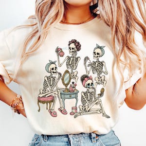 Pode incluir: Uma t-shirt de cor creme com uma impressão gráfica de quatro esqueletos a preparar-se. Os esqueletos usam acessórios de cabelo e produtos de beleza como perfume, batom e espelhos compactos em torno de uma pequena mesa de toucador.