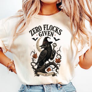 May include: A cream-colored t-shirt featuring a graphic print of a black raven wearing a witch hat, perched on a gnarled branch. The raven holds a skeleton key. The design includes a crescent moon, bats, and autumn leaves with the text ZERO FLOCKS GIVEN.