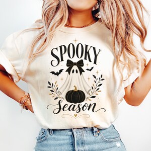 May include: A cream-colored t-shirt featuring a graphic print of a white ghost wearing a black bow, positioned above a black pumpkin. The text reads "SPOOKY" at the top and "Season" at the bottom, surrounded by small black bats, gold stars, and leafy branches.