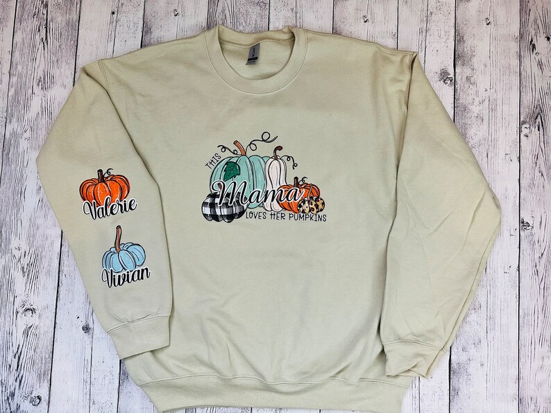 Pode incluir: Uma camisa de frio verde claro com o texto "This Mama Loves Her Pumpkins" e um gr&aacute;fico de ab&oacute;boras. As mangas t&ecirc;m os nomes "Valerie" e "Vivian" nelas.