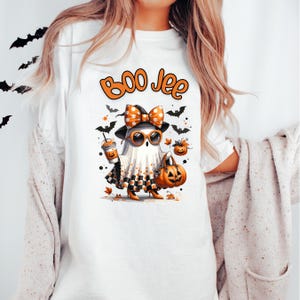 May include: White t-shirt with a Halloween-themed graphic. The design features a cartoon ghost wearing sunglasses, a bow, and holding a pumpkin bag and a coffee cup. The text "Boo Jee" is above the ghost, with bats and pumpkins scattered around.