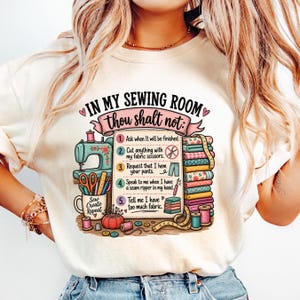 May include: A cream-colored t-shirt featuring a graphic print with the text "IN MY SEWING ROOM thou shalt not:" followed by a numbered list of five sewing rules, a teal sewing machine, fabric stacks, scissors, and various sewing supplies.