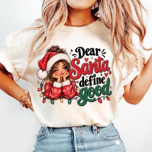 May include: A cream-colored t-shirt featuring a graphic of a person wearing a red Santa hat and a red patterned sweater. The text reads Dear Santa, define good in red and green script, surrounded by small red hearts and colorful Christmas lights.
