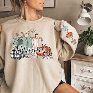 Pode incluir: Uma camisa de moletom bege com um gr&aacute;fico de ab&oacute;boras e o texto "This Mama Loves Her Pumpkins". A manga tem um remendo com tr&ecirc;s ab&oacute;boras e os nomes "Josie", "Evan" e "Cole".